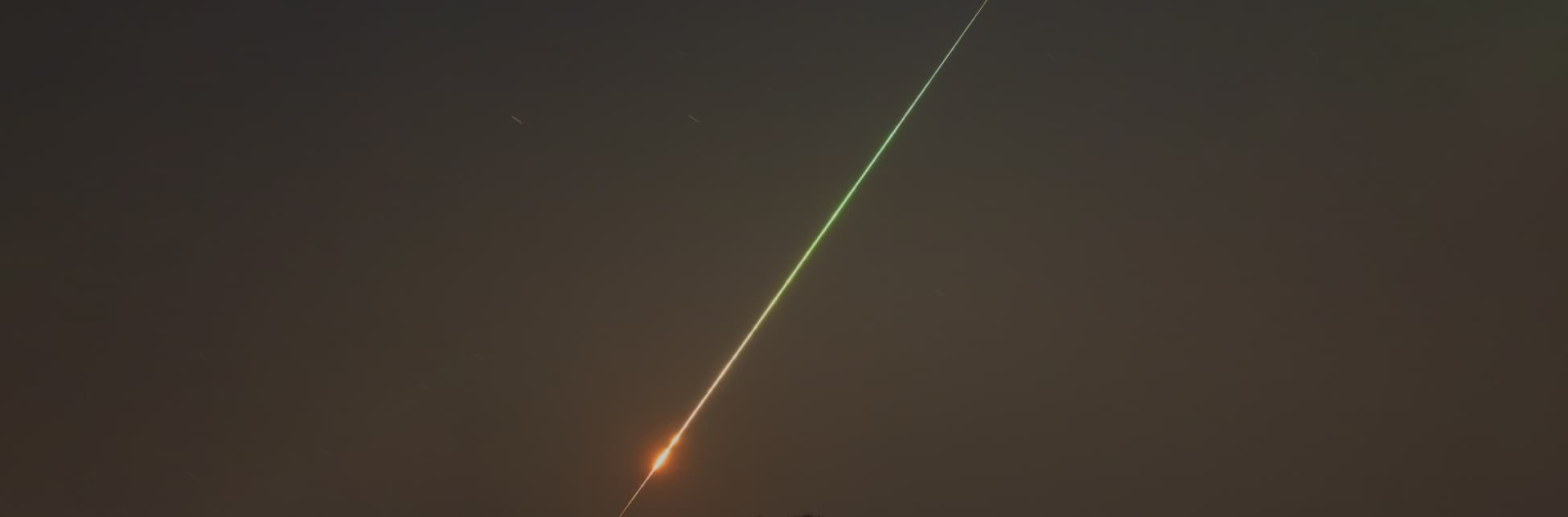 Foto. Atravesando un cielo que aún no está oscuro del todo, una línea que comienza siendo una luz amarilla y se convierte en verde. En un punto de la línea, esta se hace más gruesa como formando un pequeño círculo. 