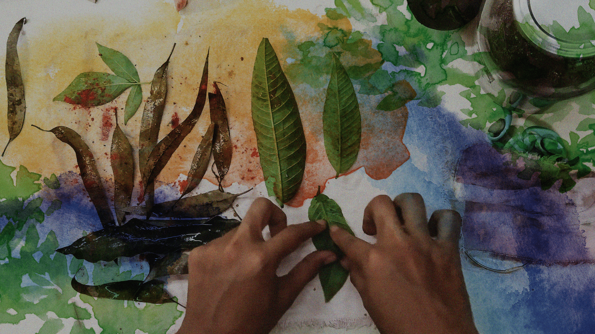 Fotografía. En el centro de la imagen dos manos están poniendo una hoja de árbol sobre un papel. El papel tiene manchas de pintura y otras hojas en desorden.