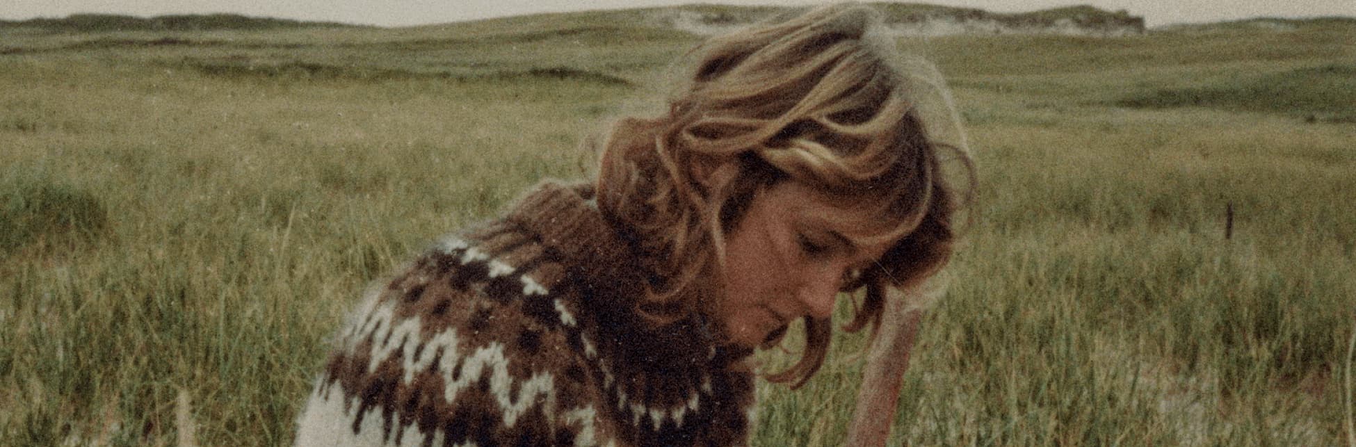 Foto. De medio cuerpo y de medio perfil, una mujer sentada. Está inclinada hacia adelante y mira un cuaderno apoyado en sus piernas. Sobre el cuaderno reposa su mano derecha, con la que sostiene un lapicero. 