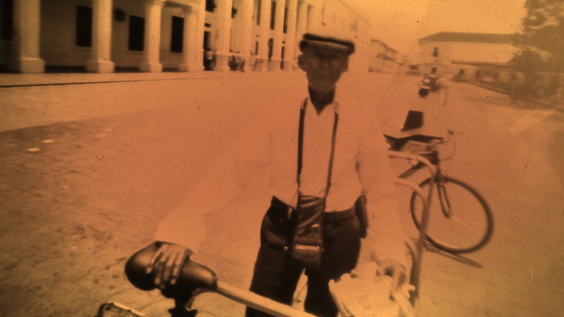 Fotografía. En primer plano, un hombre se encuentra de pie sosteniendo una bicicleta, su figura aparece desenfocada y ligeramente distorsionada. Lleva gorra, camisa clara y tirantes, y tiene un bolso colgado al frente del cuerpo. En la parte inferior izquierda se observa el sillín de otra bicicleta. Al fondo, se distingue una calle amplia con edificios de columnas a la izquierda y otras construcciones hacia la derecha, junto con algunas figuras humanas y vehículos. La imagen presenta un tono sepia uniforme y múltiples manchas y puntos dispersos en toda la superficie.