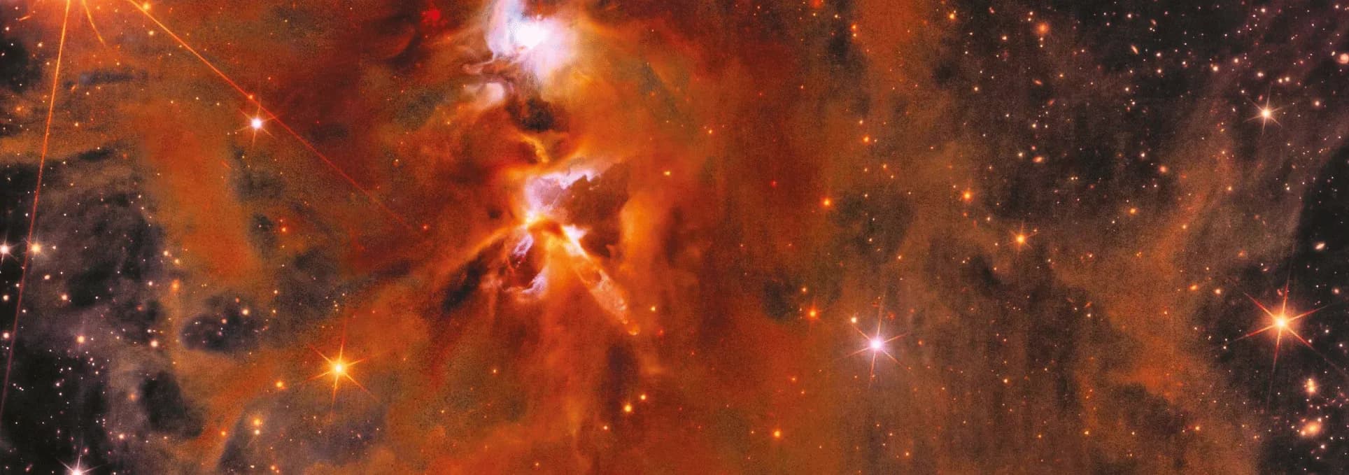 Imagen. Se observa una formación cósmica con nubes de gas y polvo interestelar en tonos rojizos y anaranjados que se mezclan con áreas más oscuras. En el centro sobresale una región brillante de color blanco con destellos luminosos, rodeada de filamentos que parecen irradiar hacia afuera. A lo largo de la imagen hay numerosas estrellas, algunas pequeñas y otras más grandes que muestran destellos de luz en forma de cruz.
