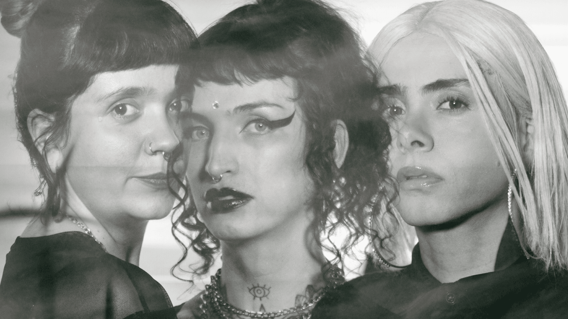 Fotografía en blanco y negro. Tres personas posan muy juntas frente a la cámara. Destacan peinados distintos, maquillaje marcado, accesorios faciales y un tatuaje de ojo en el cuello de la persona central.