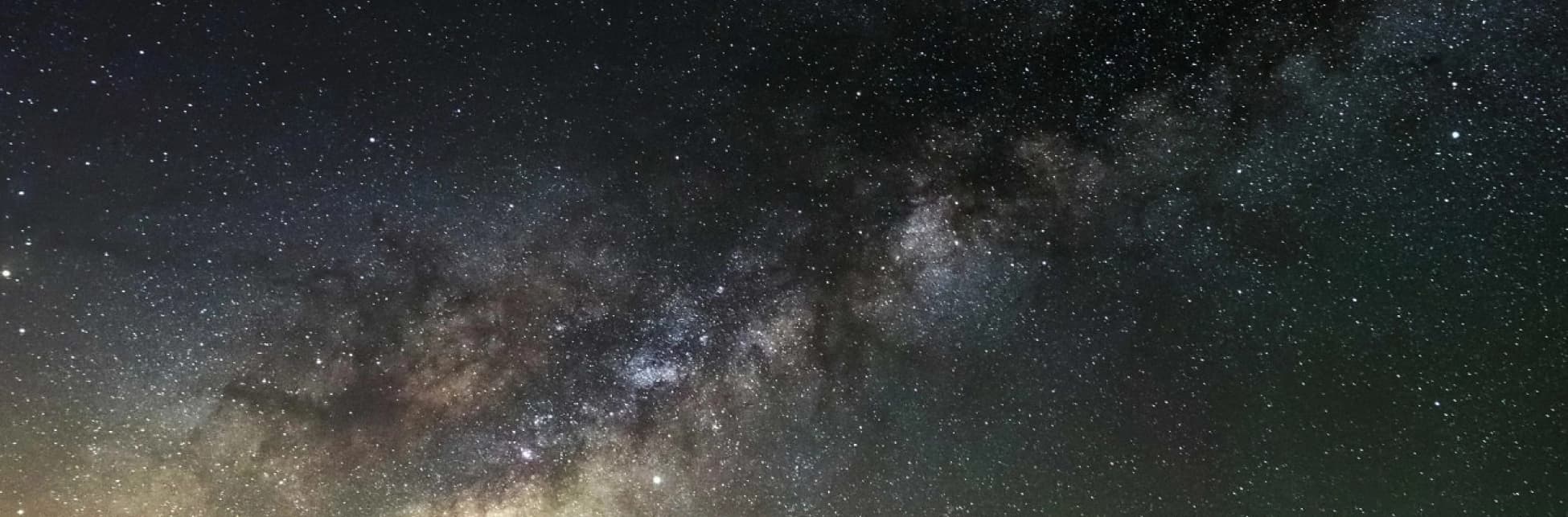 Fotografía panorámica de un cielo nocturno cubierto por numerosas estrellas. Una franja densa y luminosa de la Vía Láctea atraviesa horizontalmente la imagen, con regiones oscuras de polvo interestelar y concentraciones de estrellas visibles sobre el fondo del firmamento.