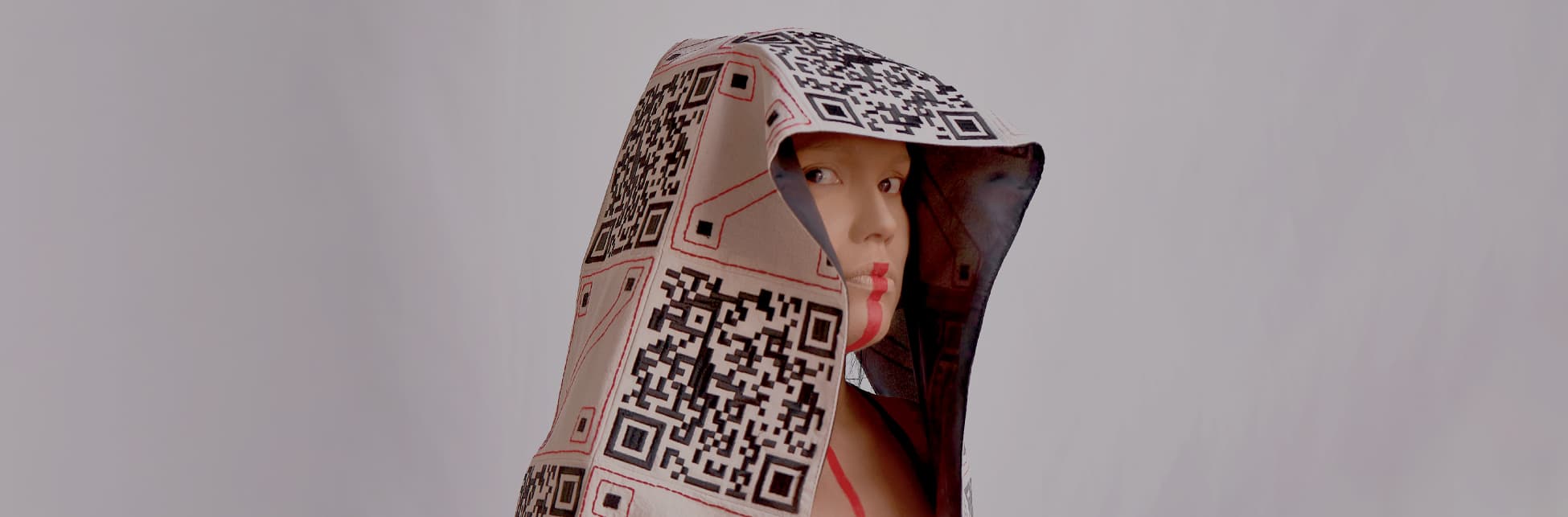 Foto. En la parte izquierda de la imagen una persona de pie, de medio cuerpo y de medio perfil. Tiene sobre su cabeza una tela bordada que cae sobre sus hombros que tiene intercalado un QR y una forma cuadrada. La persona mira directamente a quien observa la imagen y tiene una línea color rojo que comienza bajo su nariz y baja de forma vertical hasta su pecho. 