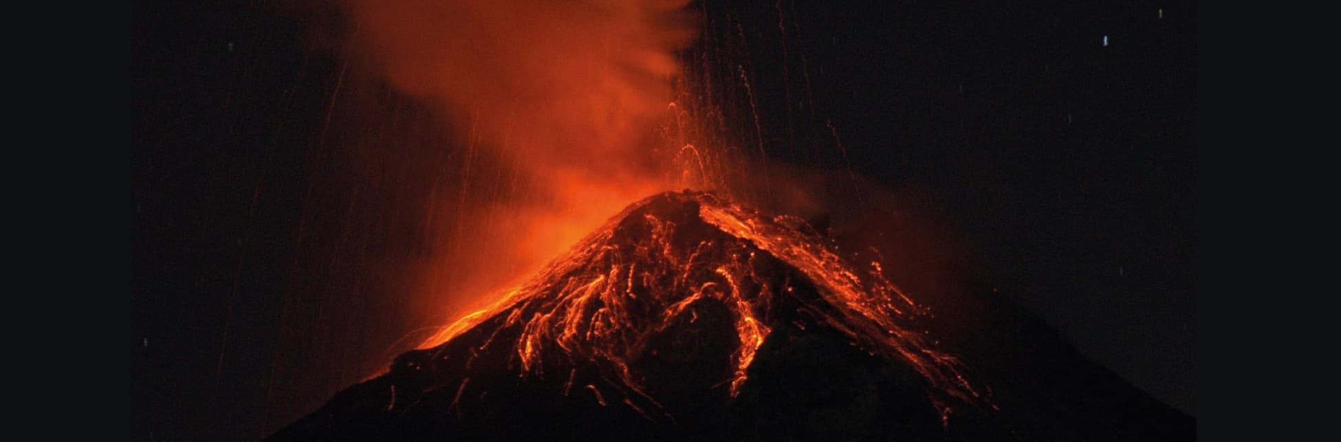 Fotografía. Imagen de un volcán en erupción durante la noche, con lava visible en la ladera y una columna de material incandescente que asciende desde el cráter.