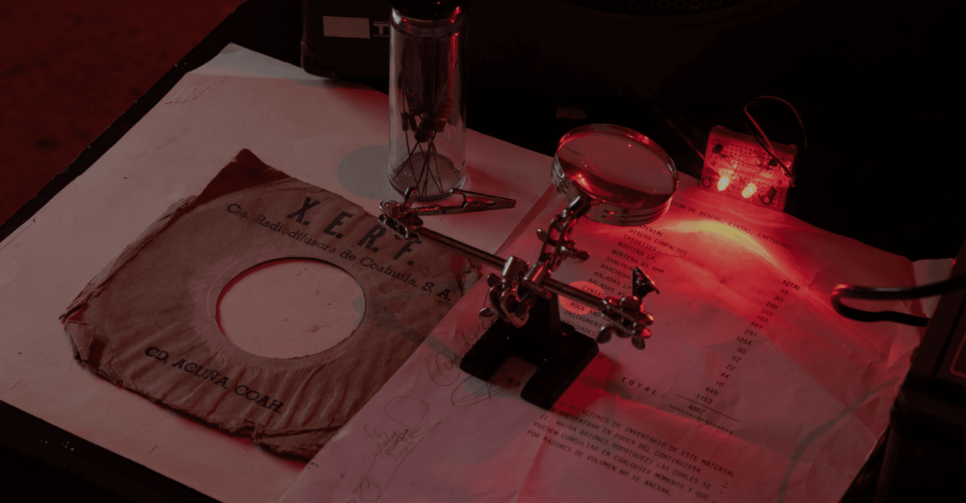 Fotografía. Sobre un escritorio, en un ambiente de investigación, la carátula de un disco de acetato y un documento son iluminados por una luz roja y observados por una lupa.