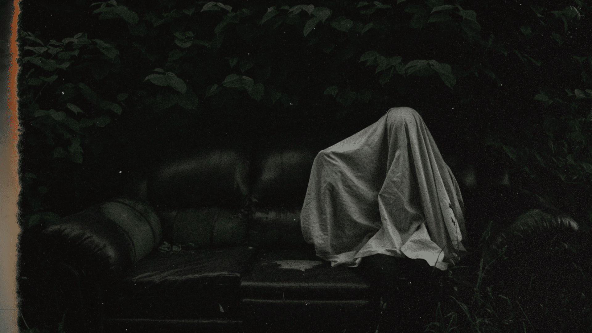 Fotografía. En un ambiente oscuro rodeado de vegetación, hay un ser cubierto por una sábana blanca sentado en un sillón, creando la apariencia de un fantasma. 