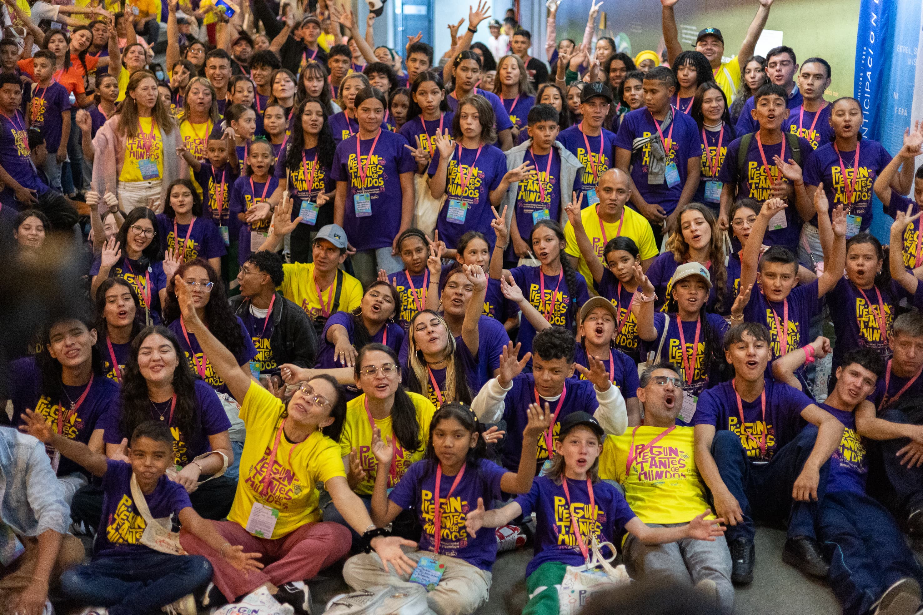 Fotografía. Grupo de niñas, niños, novenes y adultos, con expresión de alegría. Unos levanta sus manos, otros sonríen. Llevan puesto camisas color morado y amarillo que tiene estampada la frase Preguntantes de mundos.
