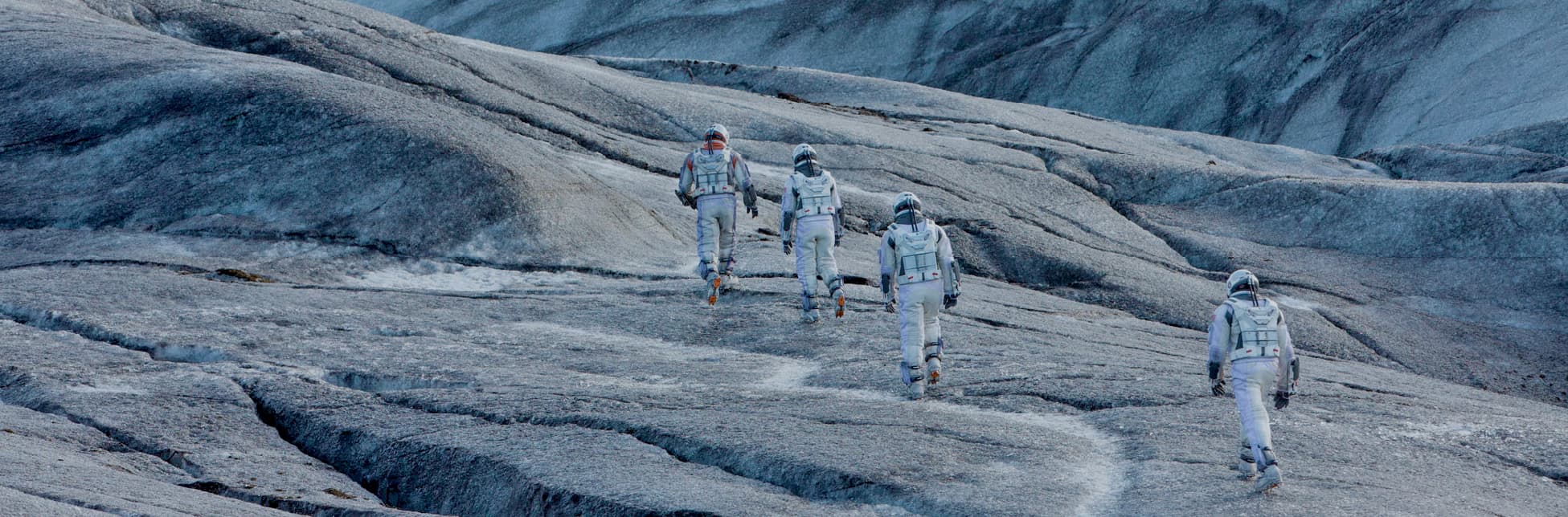 Fotograma de la película Interstelar. Sobre un paisaje rocoso de tonalidades azul grisáceo, cinco personas con trajes espaciales caminan hacia una formación rocosa. 