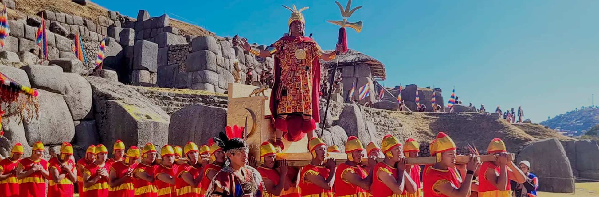 Foto. En la parte inferior de la imagen varias personas vestidas con trajes indígenas color rojo y dorado cargan una silla dorada sobre la que va una persona de pie con un vestido en tonos rojos, una corona dorada con varias puntas y un cetro dorado, al fondo varias construcciones en roca.