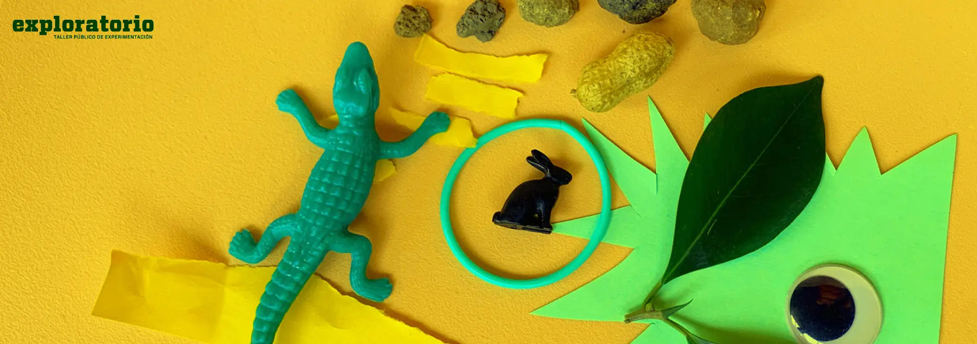 Fotografía. Sobre una superficie amarilla se encuentran diversos objetos: un cocodrilo de plástico verde, una figura negra de conejo dentro de un aro turquesa, una hoja verde natural sobre una cartulina recortada en picos, un ojo móvil, piedras de distintos tamaños y papeles rasgados en tonos amarillos.