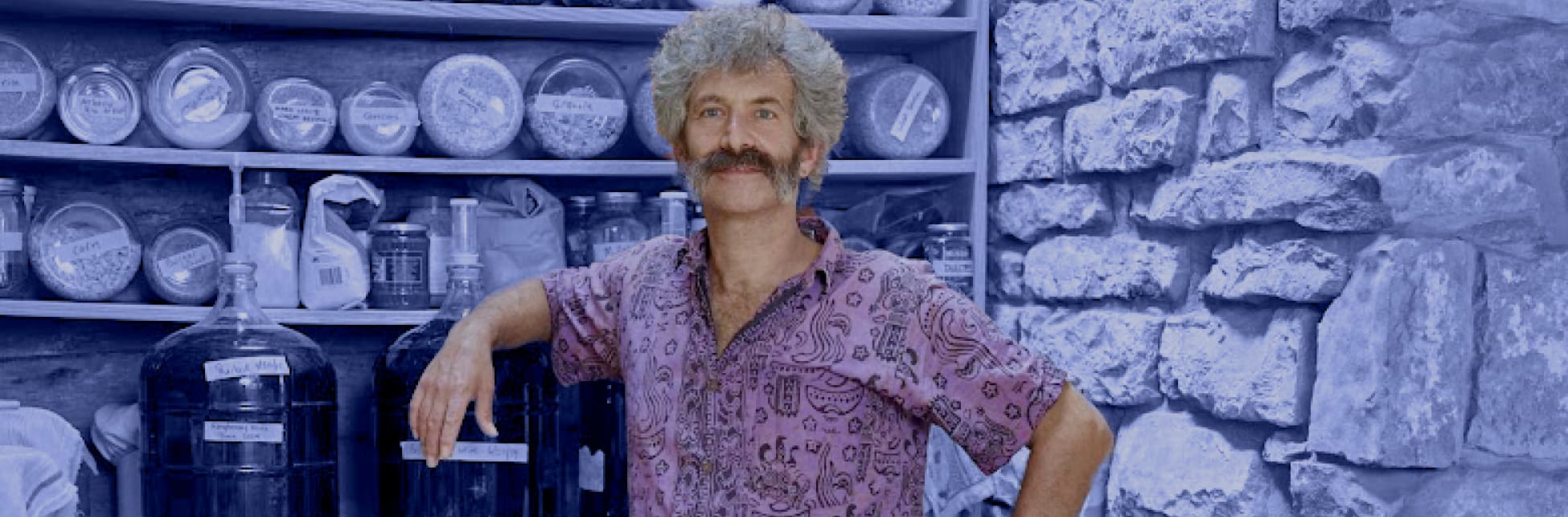 Fotografía intervenida. En el fondo, con un uniforme color azul, se ven muros, estanterías y frascos, y en el primer plano un hombre de frente, con camiseta lila ilustrada, bigote abundante y cabello blanco. 