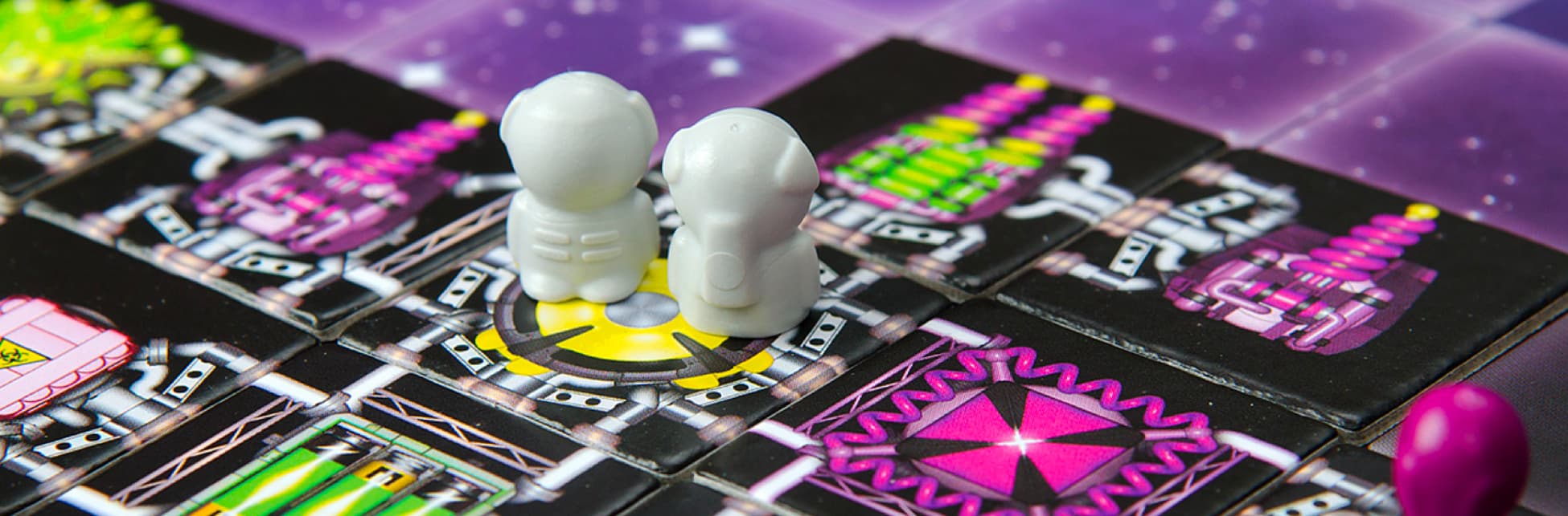 Fotografía. Tablero del juego Galaxy Trucker adornado con colores vibrantes y diversas formas geométricas, sobre el tablero dos pequeñas figuras de plástico color gris. 