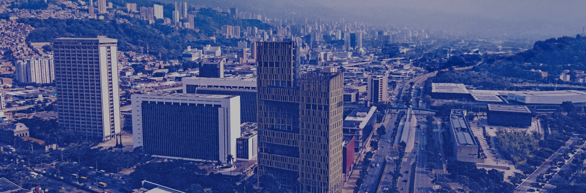 Fotografía. En la parte inferior, se observa una panorámica urbana con edificios modernos, una avenida principal y colinas al fondo. En la parte superior, el cielo está estrellado con una vista de la Vía Láctea. 