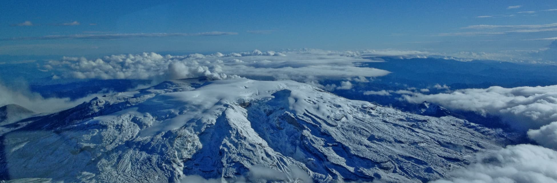 Foto. En el centro de la imagen una montaña cubierta de nieve vista desde arriba, la rodean varias nubes.