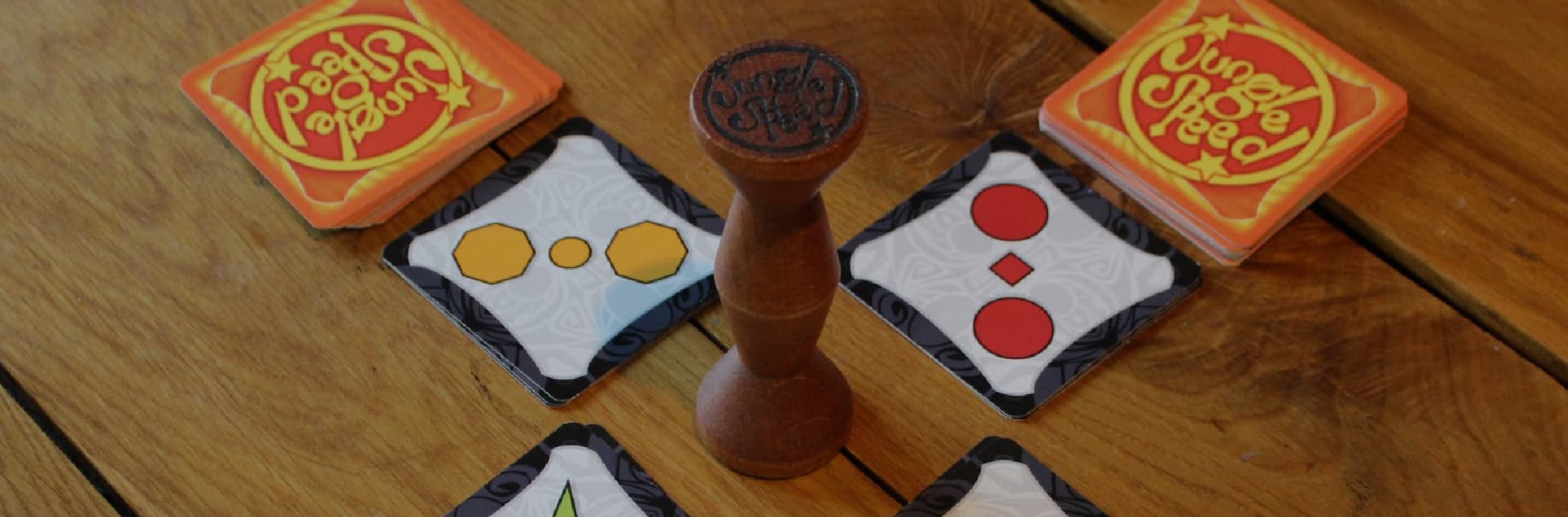 Ilustración. Sobre una mesa de madera se encuentran varias cartas del juego de mesa Jungle Speed, con formas geométricas de colores y un tótem de madera en el centro con la inscripción "Jungle Speed" en la parte superior. 