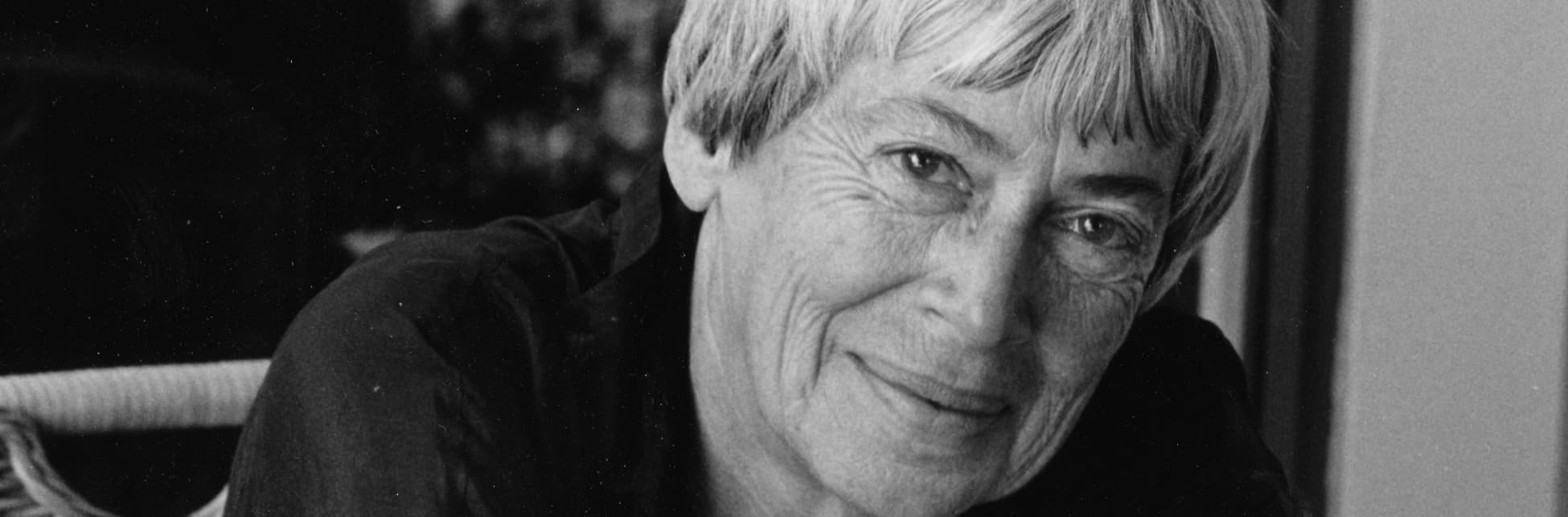 Fotografía. En blanco y negro del rostro de Ursula K. Le Guin, con el cabello corto y una sonrisa gentil. Sus ojos expresivos y sus cejas poco marcadas. 

