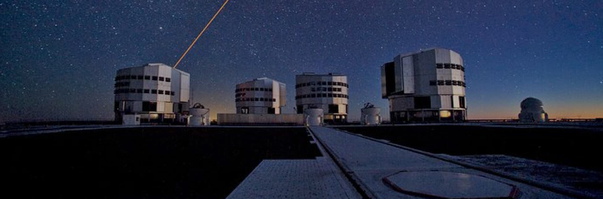 Fotografía. Cuatro edificios en forma similar a un cilindro, de uno de ellos sale una línea amarilla hacia el cielo, detrás de los edificios un cielo nocturno estrellado.
