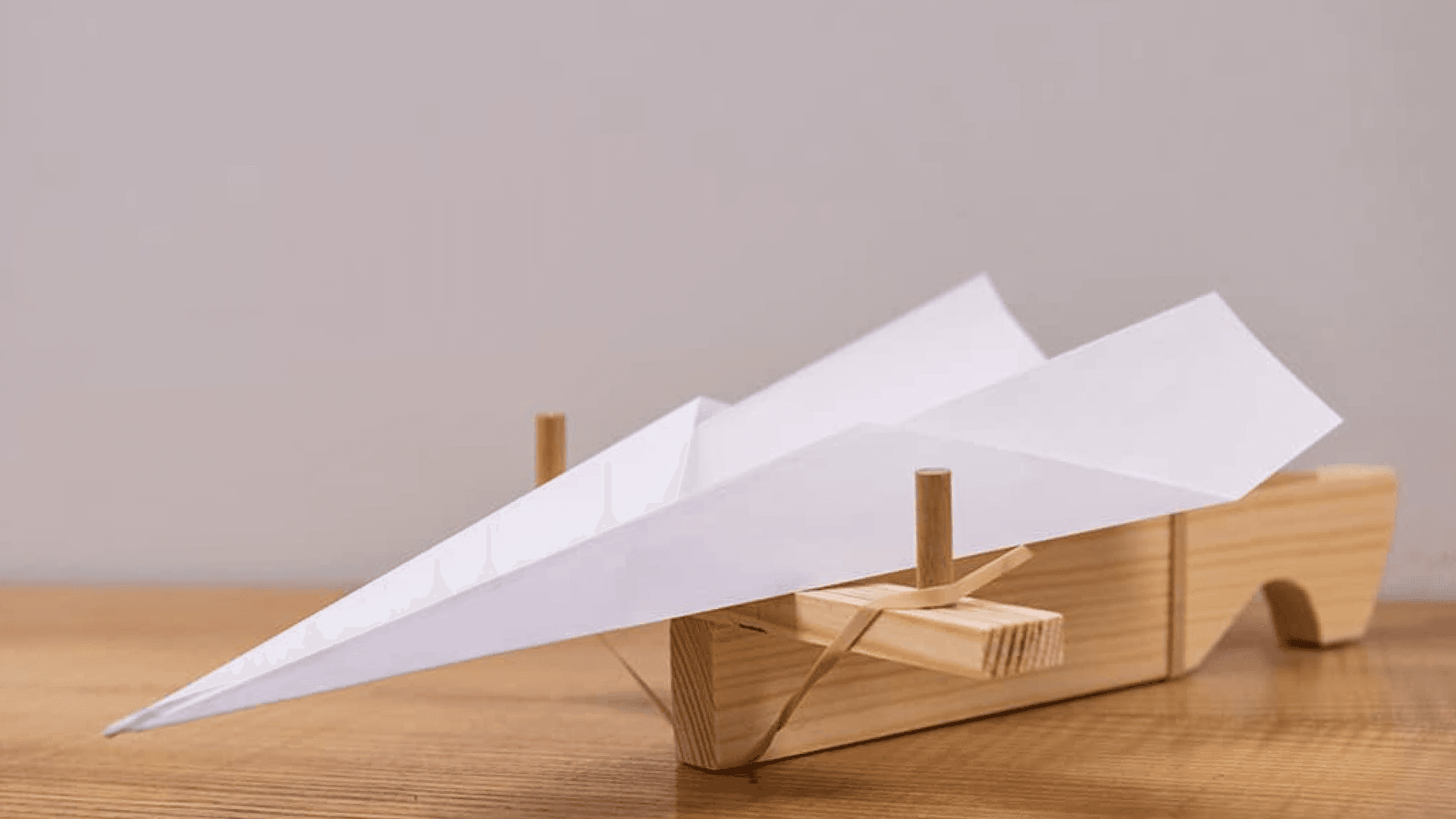 Fotografía. Sobre un fondo gris claro hay un avión de papel blanco posado sobre una plataforma de lanzamiento hecha con madera.