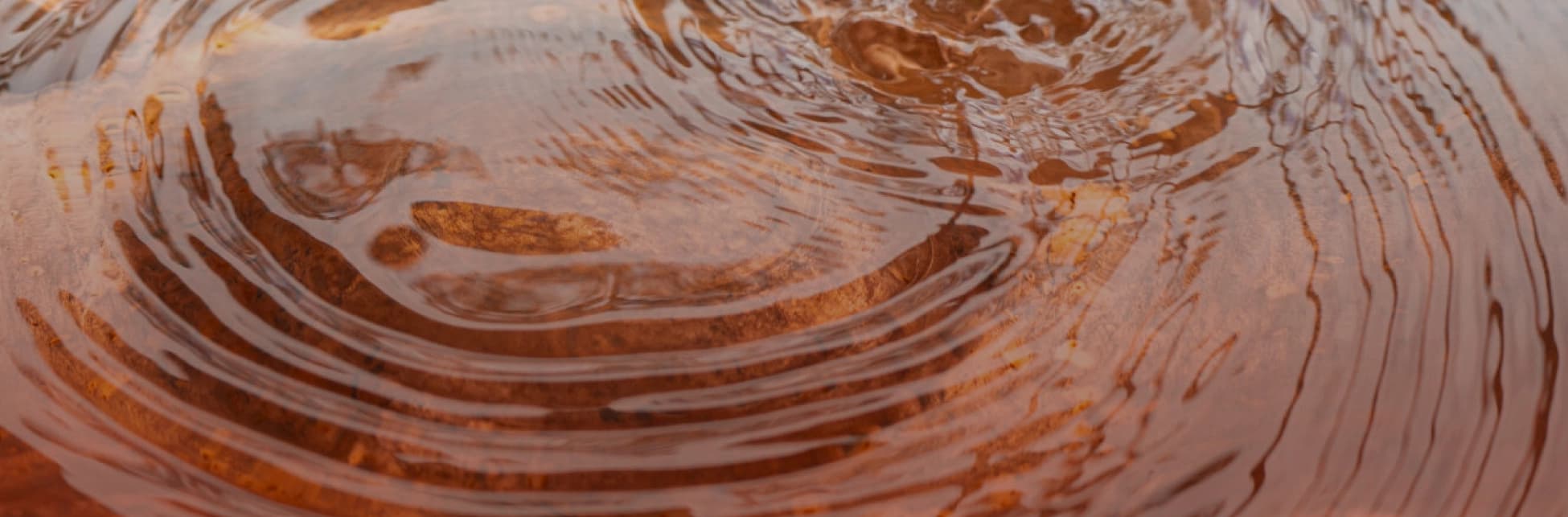 Fotografía. En primer plano se observa la superficie de un líquido rojizo con ondas concéntricas que se propagan sobre una base de piedra sumergida. Las ondulaciones generan reflejos distorsionados de estructuras cercanas, posiblemente metálicas, visibles en la parte superior derech