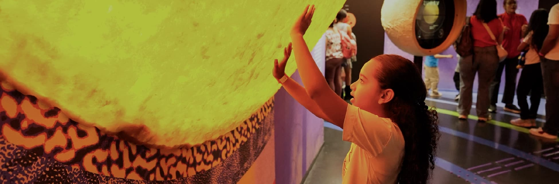 Fotografía de una niña que toca con ambas manos una gran superficie esférica iluminada, dentro de una sala con otras personas y elementos expositivos.