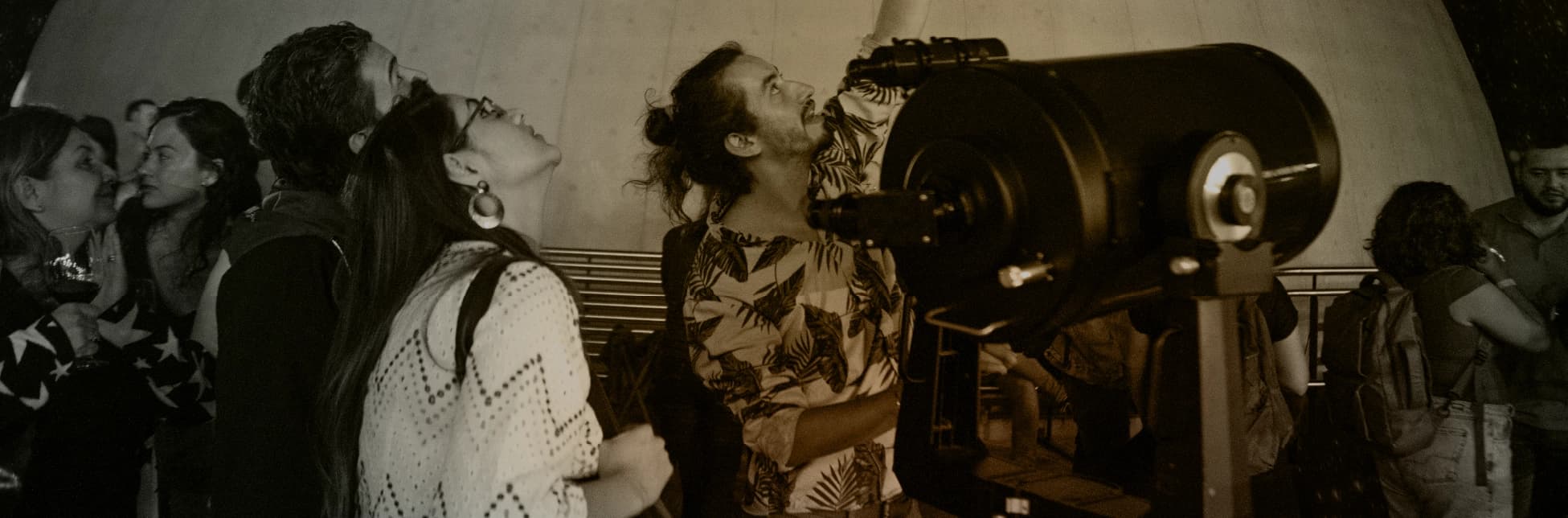 Fotografía. En una terraza de observación, varias personas miran hacia el cielo junto a un telescopio; una de ellas señala hacia arriba.