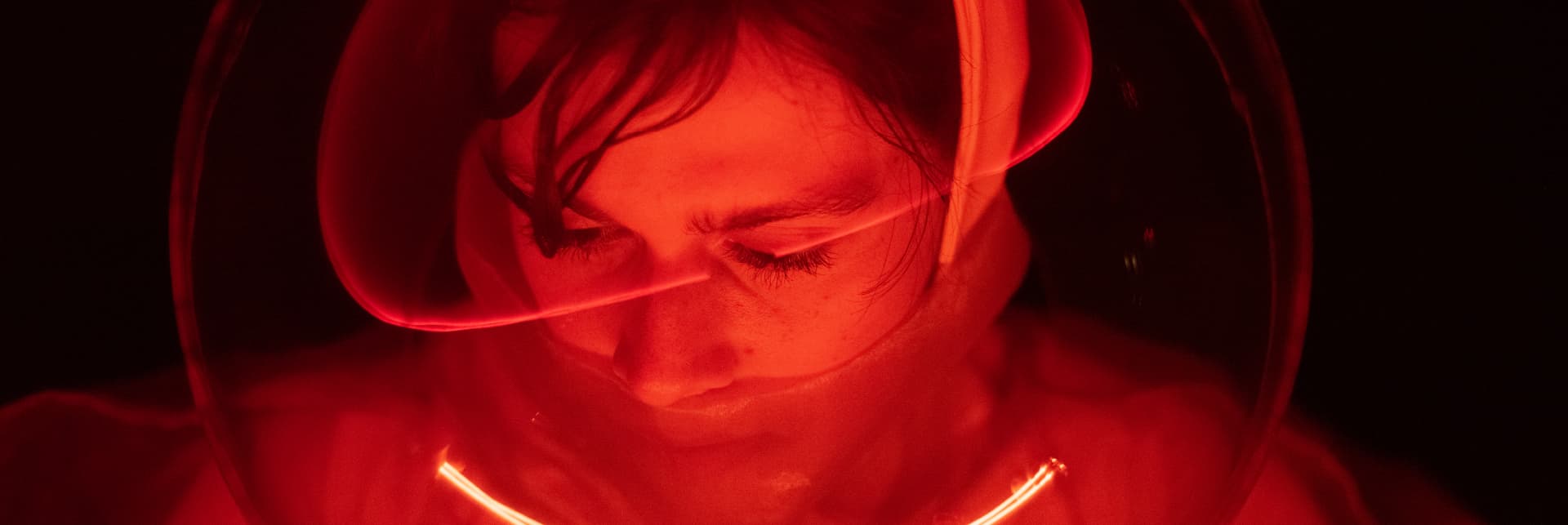 Fotografía. Iluminada en tonos rojizos aparece una mujer usando un casco espacial, con la mirada levemente inclinada hacia abajo.
Sobre un fondo negro, un texto dice: "Coctel de apertura de la Exposición El Espacio que somos. Arte y ciencia confrontan el machismo, el especismo, el progreso y la violencia. Santiago Herrera, Paulina Escobar, Harold Ortiz, Juliana Gómez. Cuatro residencias artísticas. Planetario de Medellín. Artes + Ciencias + Tecnologías + Sociedad. 2022-2023".