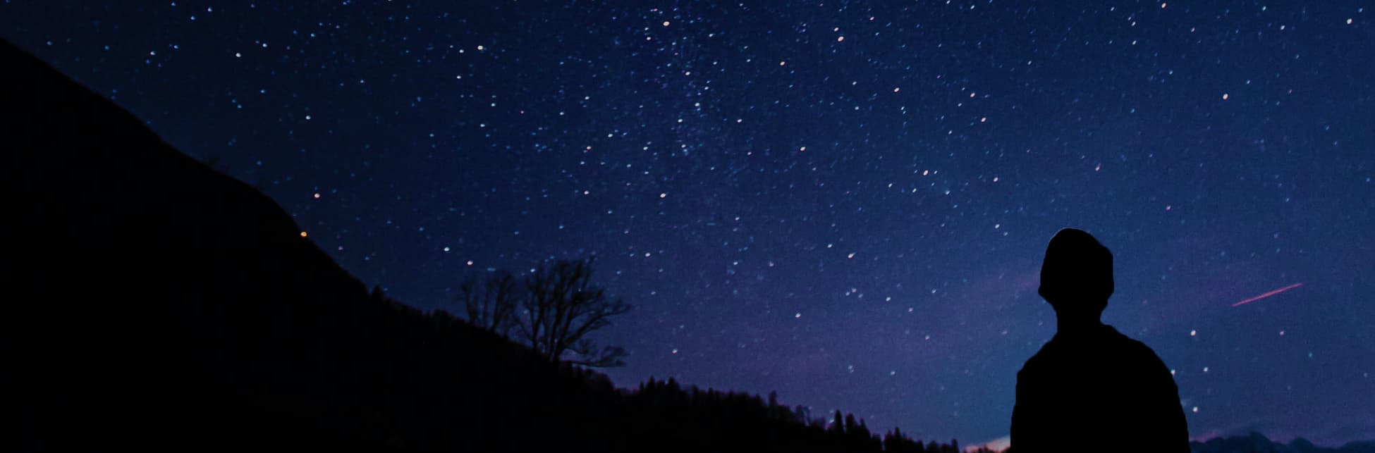 Foto. Sobre un fondo estrellado aparece en la parte inferior de la imagen la silueta de una pequeña montaña y una persona.