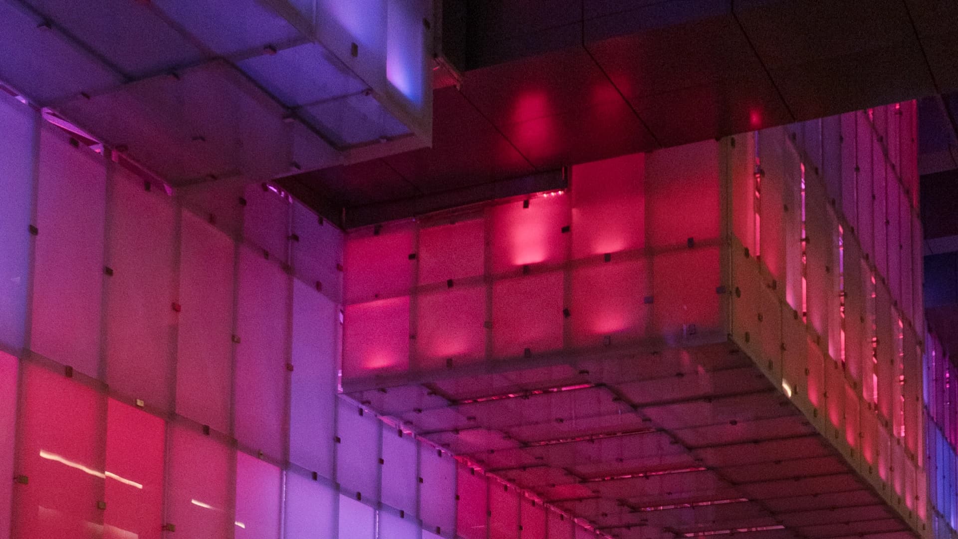 Fotografía. Interior de un espacio arquitectónico con paredes y techo formados por paneles rectangulares translúcidos y estructuras modulares. La iluminación en tonos rosados, violetas y azules se proyecta sobre las superficies y crea variaciones de luz y sombra. A la derecha, una estructura elevada se prolonga hacia el fondo y forma una sucesión de paneles verticales. En la parte superior se observa un techo oscuro que contrasta con las zonas iluminadas.