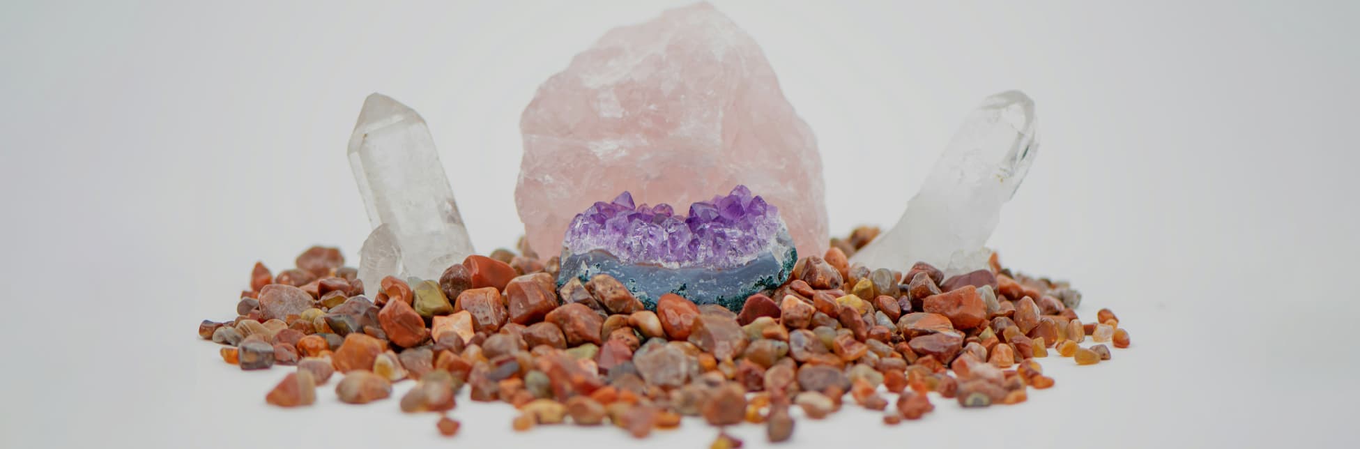 Ilustración. Sobre un fondo claro se observa una composición de minerales: al centro hay una gran roca rosada, delante de ella un fragmento de amatista morada y a cada lado dos cristales transparentes; todas las piezas reposan sobre numerosas piedras pequeñas en tonos cafés y rojizos.