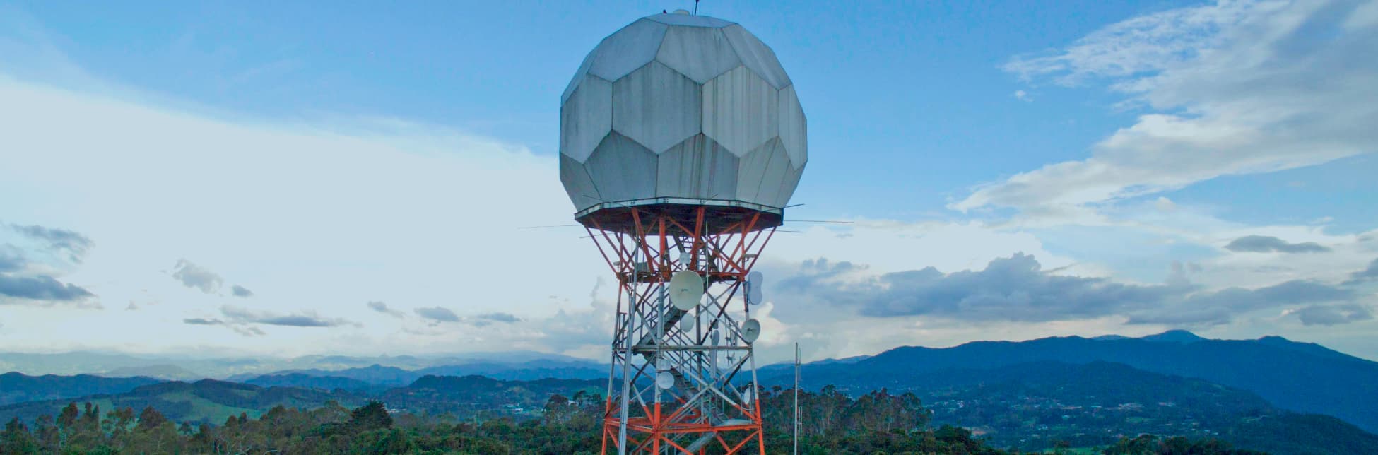 Fotografía. Sobre un fondo de cielo azul con nubes, se ve una gran estructura metálica roja que sostiene una cúpula geométrica blanca similar a un radar; alrededor hay antenas y, al fondo, montañas.