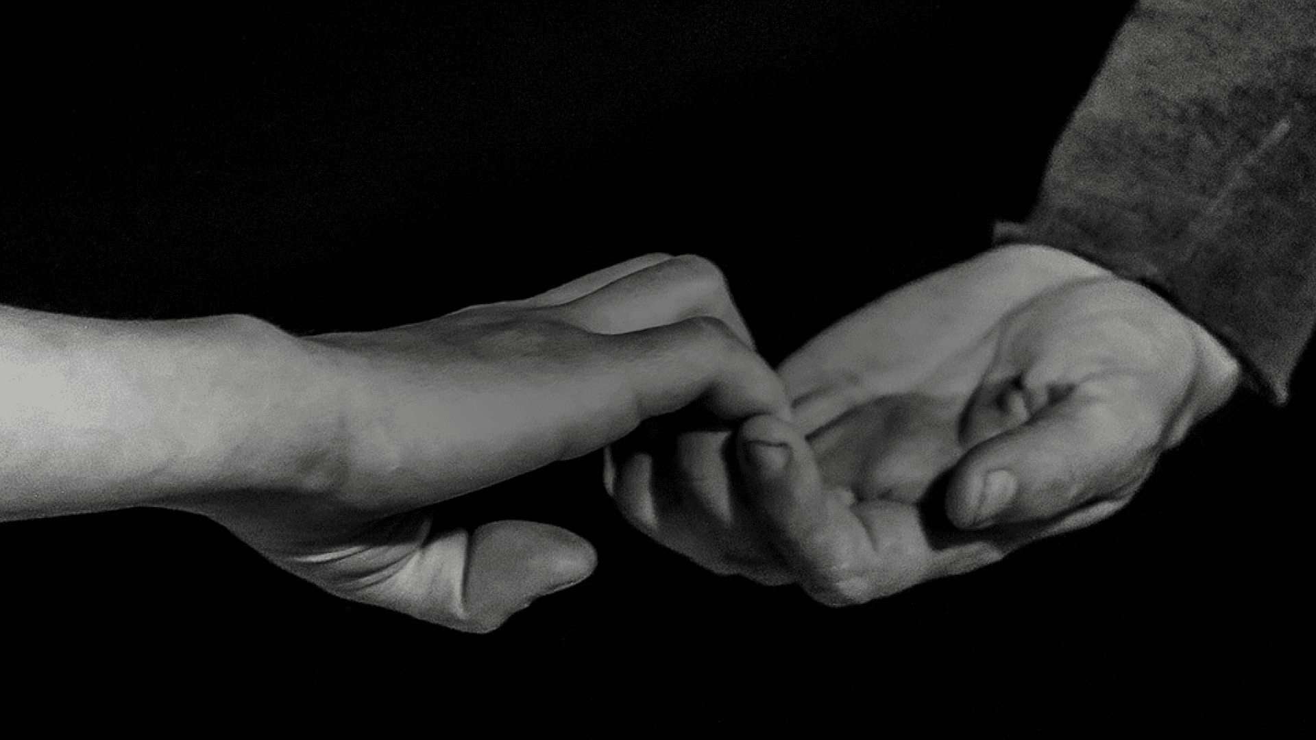 Fotografía en blanco y negro. Sobre un fondo negro, hay dos manos sujetándose con los dedos extendidos, evocando un gesto de conexión.