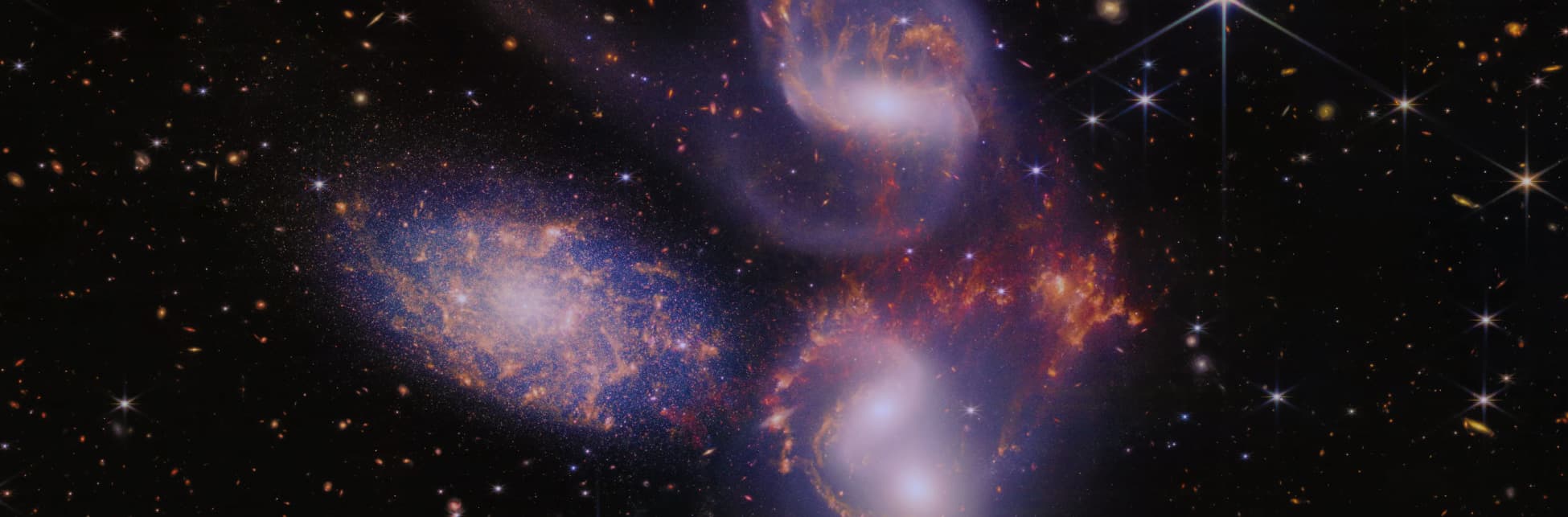 Imagen digital. Cuatro puntos de luz rodeados por una sombra blanca, son galaxias, a su alrededor destellos y brillos de colores naranja, azul y blanco.