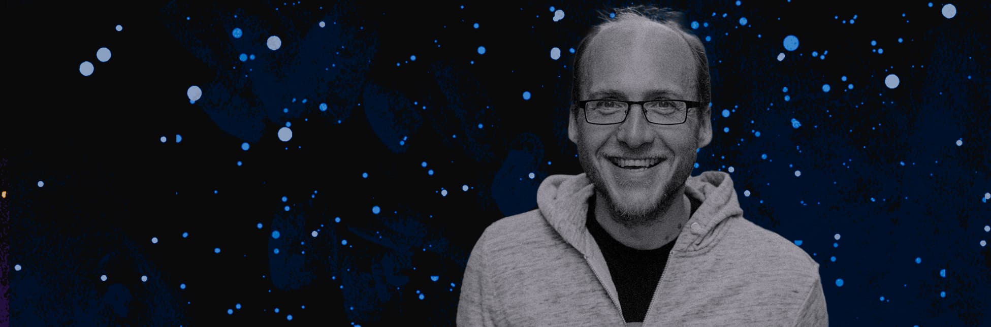 Fotografía intervenida. En la parte derecha de la imagen un hombre sonriente con gafas y sudadera clara en primer plano sobre un fondo oscuro estrellado.