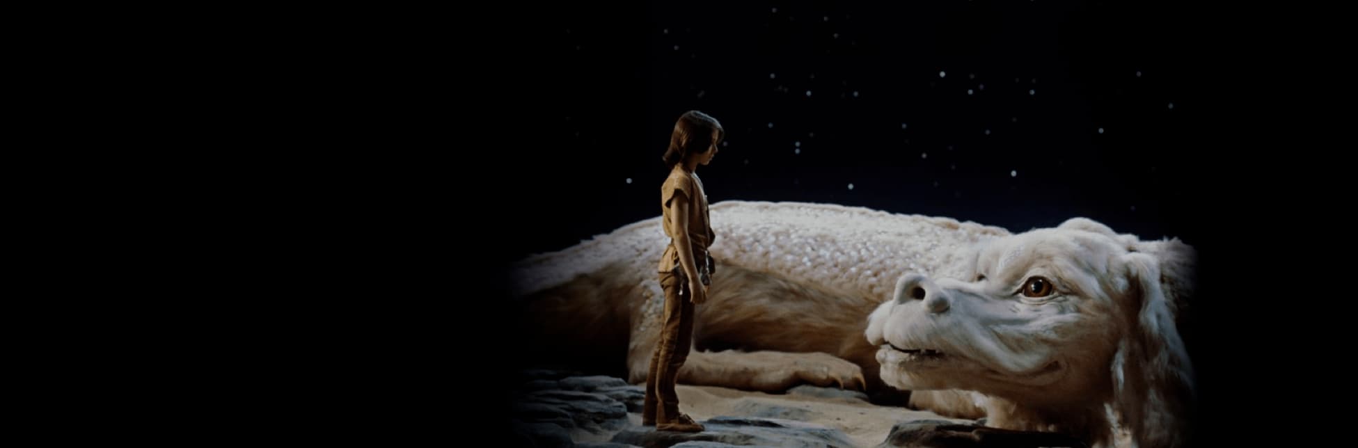 Foto. En el centro de la imagen, un niño frente a un dragón blanco de gran tamaño con expresión amable, sobre un fondo de cielo estrellado.
