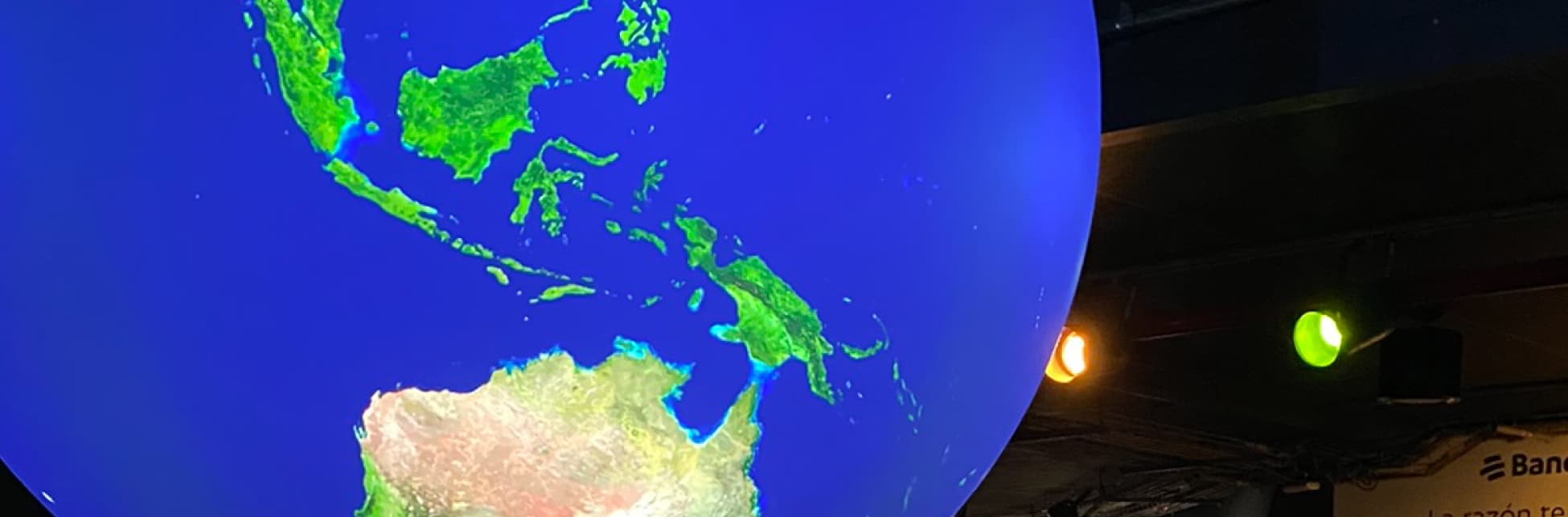Foto. En la parte izquierda de la imagen una esfera con el globo terráqueo, una esfera azul y verde que representa el planeta Tierra. Varias personas miran con atención.