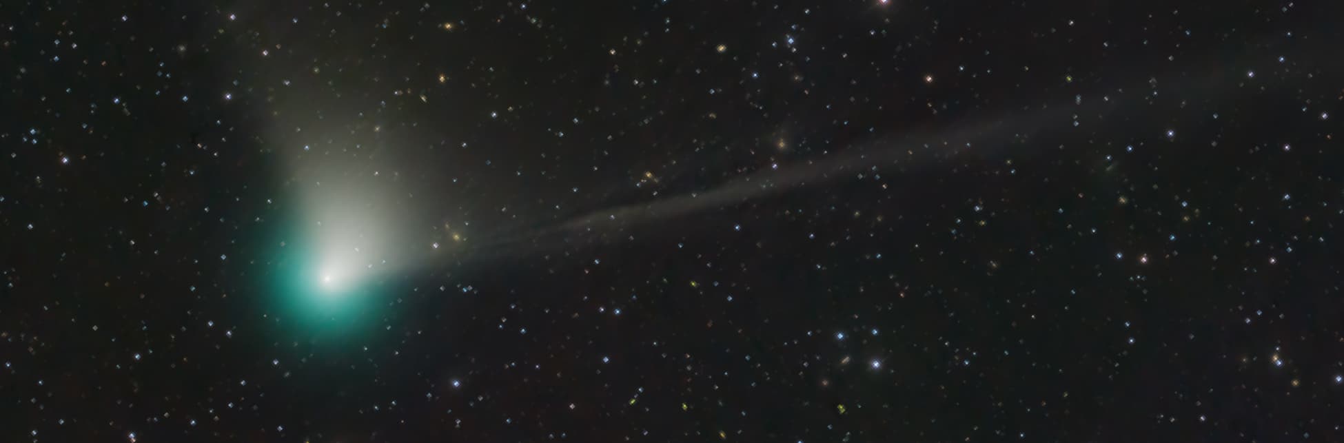 Foto horizontal. Sobre un fondo estrellado aparece en la parte inferior izquierda de la imagen un comenta: un punto que irradia una luz verdosa y de él se desprende un halo de luz blanca hacia arriba.
