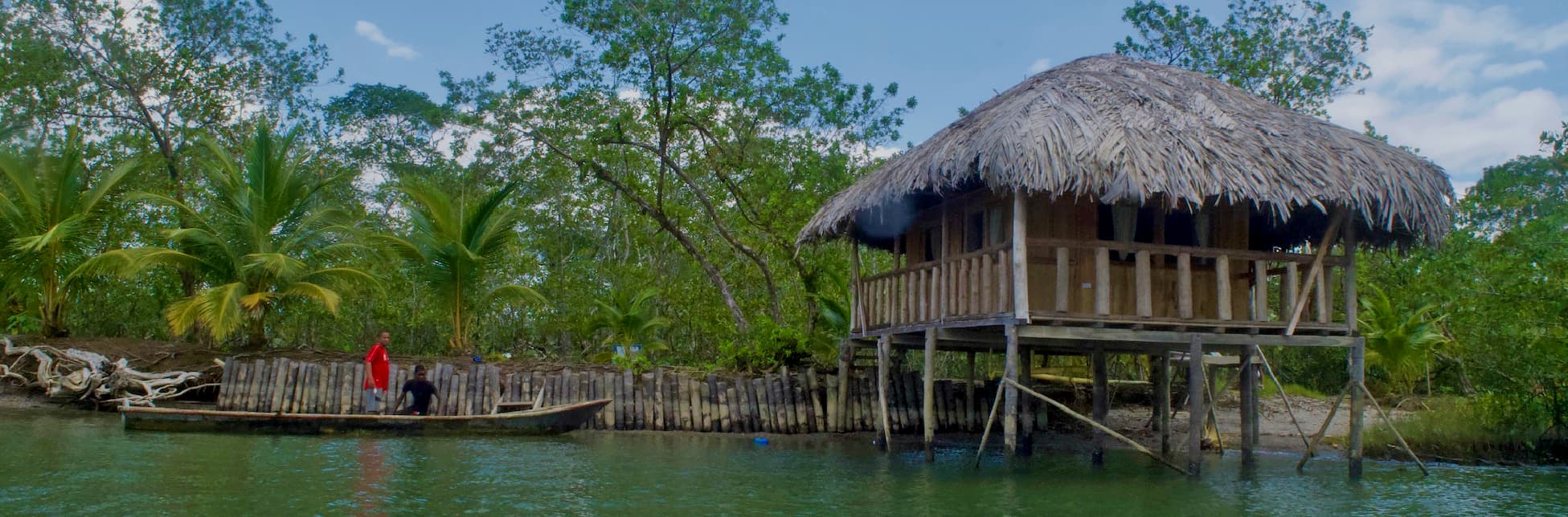 Fotografía. En la parte inferior se observa una casa de madera elevada sobre pilotes junto al agua, con techo de palma, rodeada de vegetación y árboles; a la izquierda hay un muelle rústico con dos personas y el agua ocupa el primer plano.