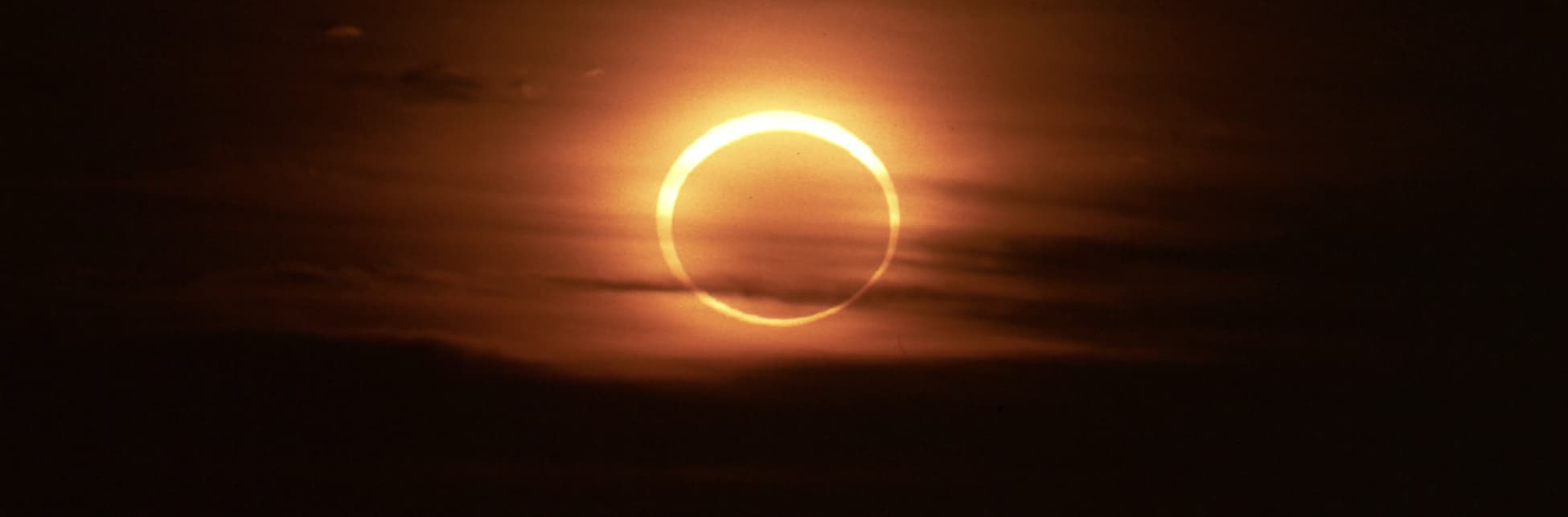 Foto. En la parte superior de la imagen un anillo de luz al fondo varias nubes en tonos naranjas.