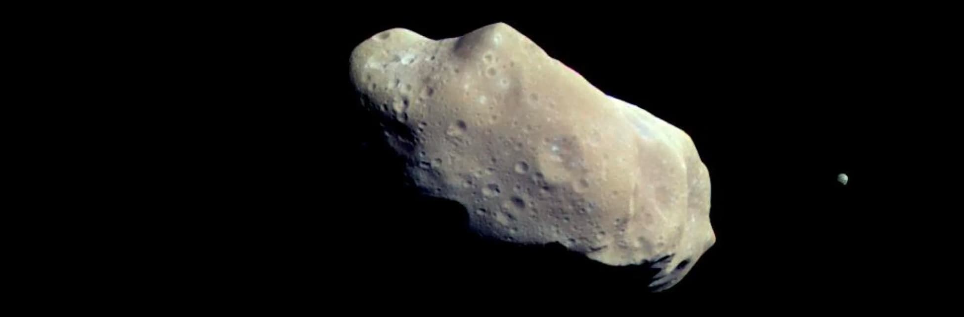 Fotografía. Sobre un fondo negro se ve una roca espacial color crema, de superficie y forma irregular y con cráteres.