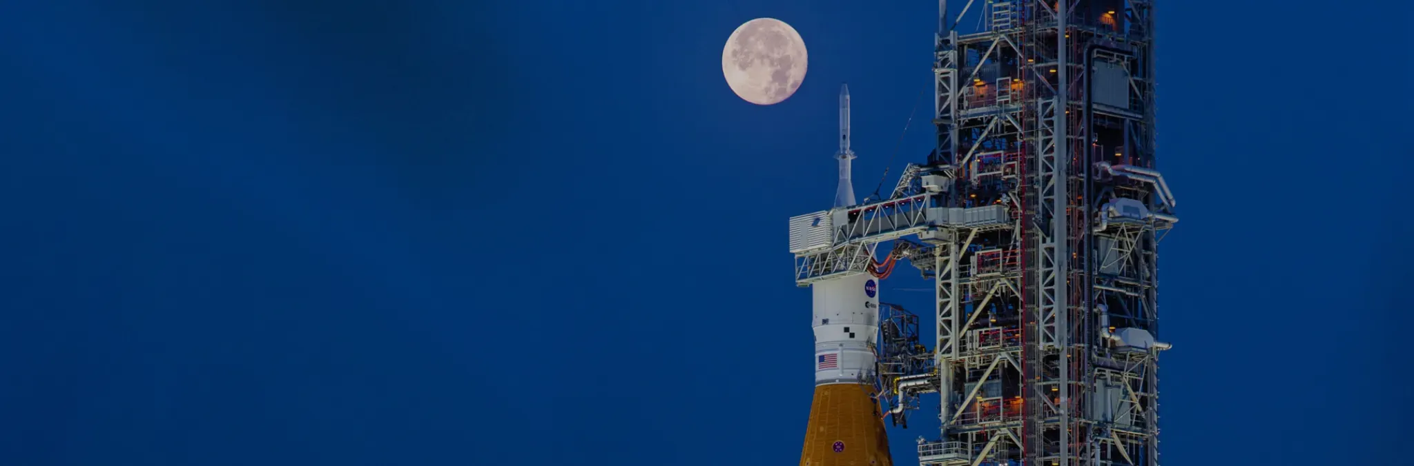 Fotografía. Sobre un fondo azul oscuro aparece, a la derecha, una estructura metálica de lanzamiento con un cohete en posición vertical y, a su izquierda, una luna llena.