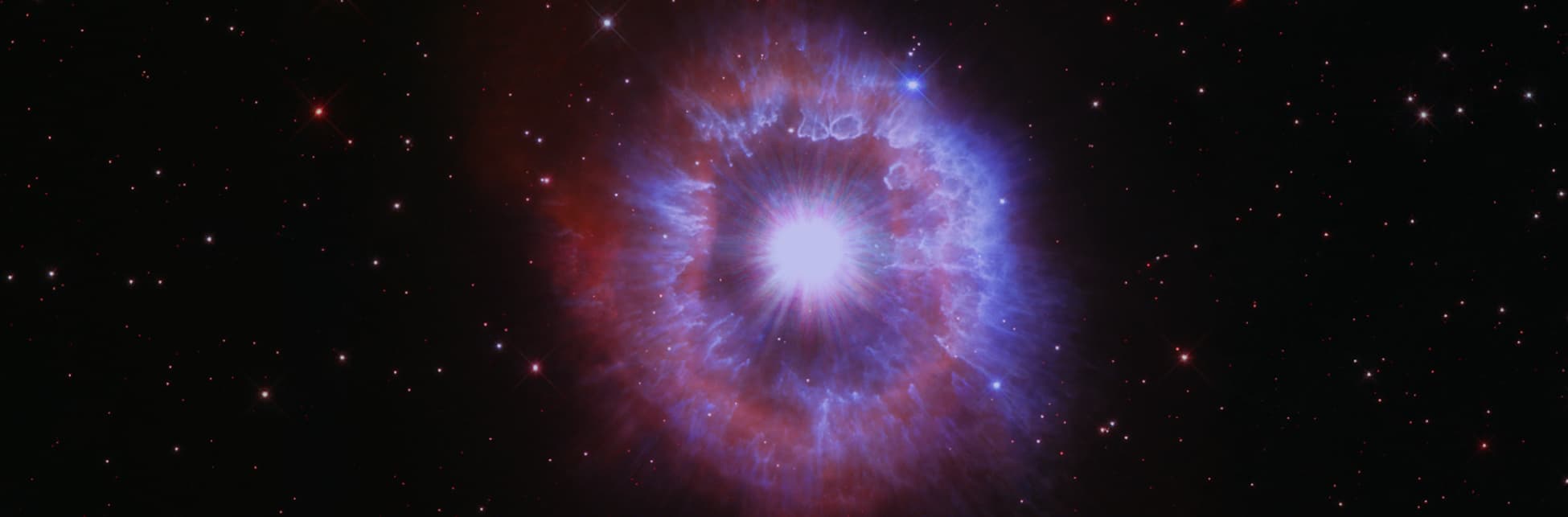 Astrofotografía. Sobre un fondo estrellado aparece un punto muy brillante del que salen rayos de luz y se van haciendo más grandes convirtiéndose en una gran mancha de tonos rojos y azules.
