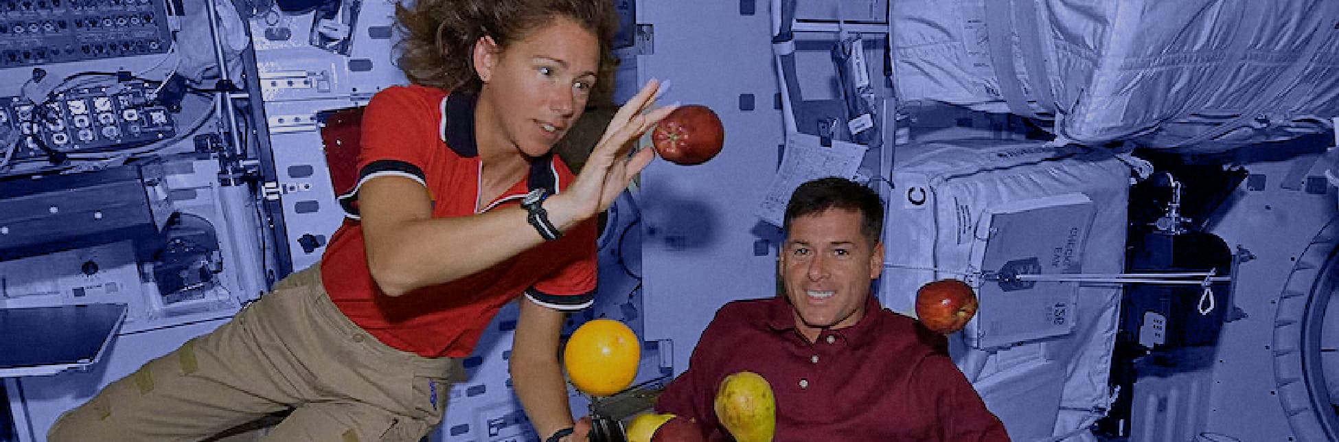 Fotografía. En el interior de una nave espacial con paredes cubiertas de paneles y dispositivos tecnológicos, flotan varias frutas, entre ellas manzanas, peras, tomates y un durazno. A la izquierda, una mujer con cabello rizado y camiseta roja con detalles azules y blancos extiende su brazo hacia un tomate. A la derecha, un hombre con camisa tipo polo de color vino tinto sonríe mientras observa las frutas flotando frente a él.