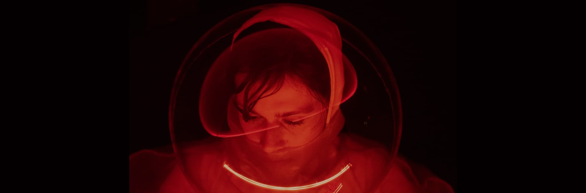 Foto horizontal. En el centro de la imagen una mujer en primer plano en tonos rojos y negro. Tiene la cabeza ligeramente inclinada hacia arriba al igual que sus ojos. Frente a su rostro parece haber una superficie transparente que refleja una luz que atraviesa en diagonal la imagen.