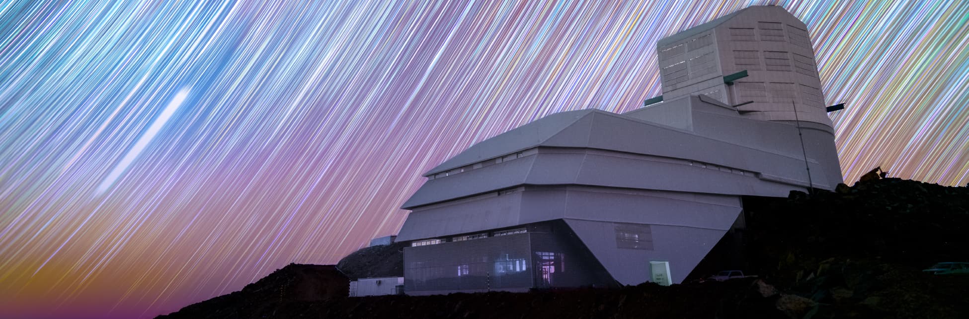 Foto. En la parte inferior de la imagen un observatorio, un edificio rectangular, en la parte delantera tiene un techo por capas, una un poco más atrás que la otra, y en la parte trasera se eleva una sección rectangular hacia arriba. Arriba, en el cielo, líneas diagonales blancas sobre un fondo rosado y azul.