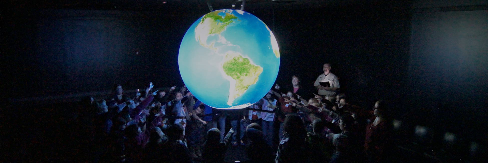 Foto. En el centro de la imagen el planeta Tierra muy brillante, una esfera azul con varias porciones de tierra color verde y café. Al rededor varias niñas y niños apuntan a la esfera.