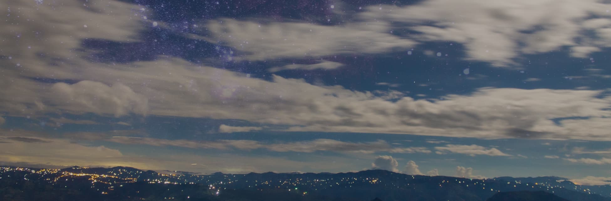 Fotografía de paisaje nocturno. En la parte inferior, se observa una vista de montañas con luces de un pueblo que brillan en la oscuridad. El cielo está parcialmente cubierto por nubes, permite ver algunas áreas despejadas que muestran estrellas y detalles de la Vía Láctea en tonos violetas y azules.