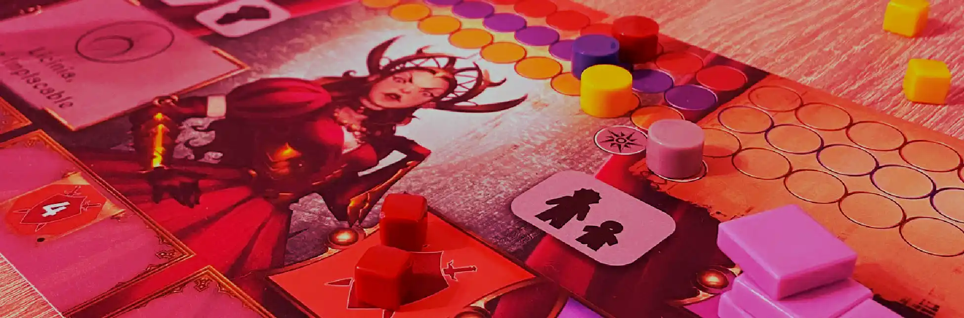 Fotografía. Sobre un fondo rojizo y rosado, se ve una mesa con un juego de mesa desplegado, fichas redondas de varios colores, cartas y figuras pequeñas.
