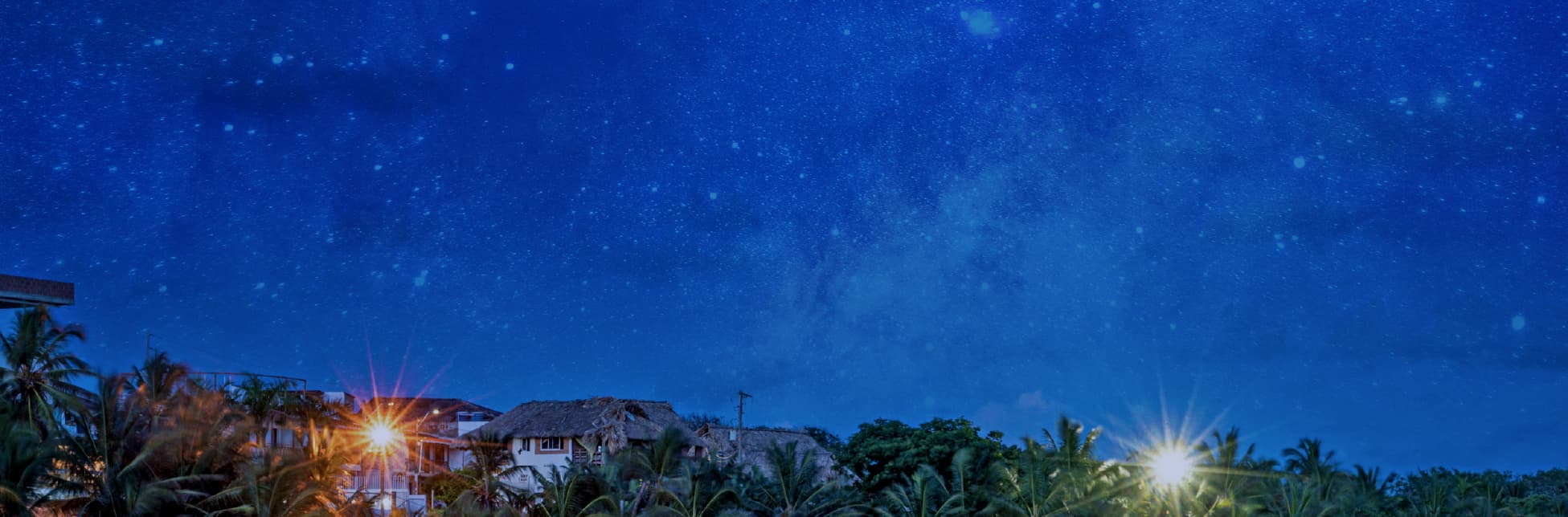 Fotografía. En la parte inferior se observa una playa al atardecer con el reflejo del cielo sobre el agua, palmeras iluminadas y casas al fondo. En la parte superior, el cielo estrellado.