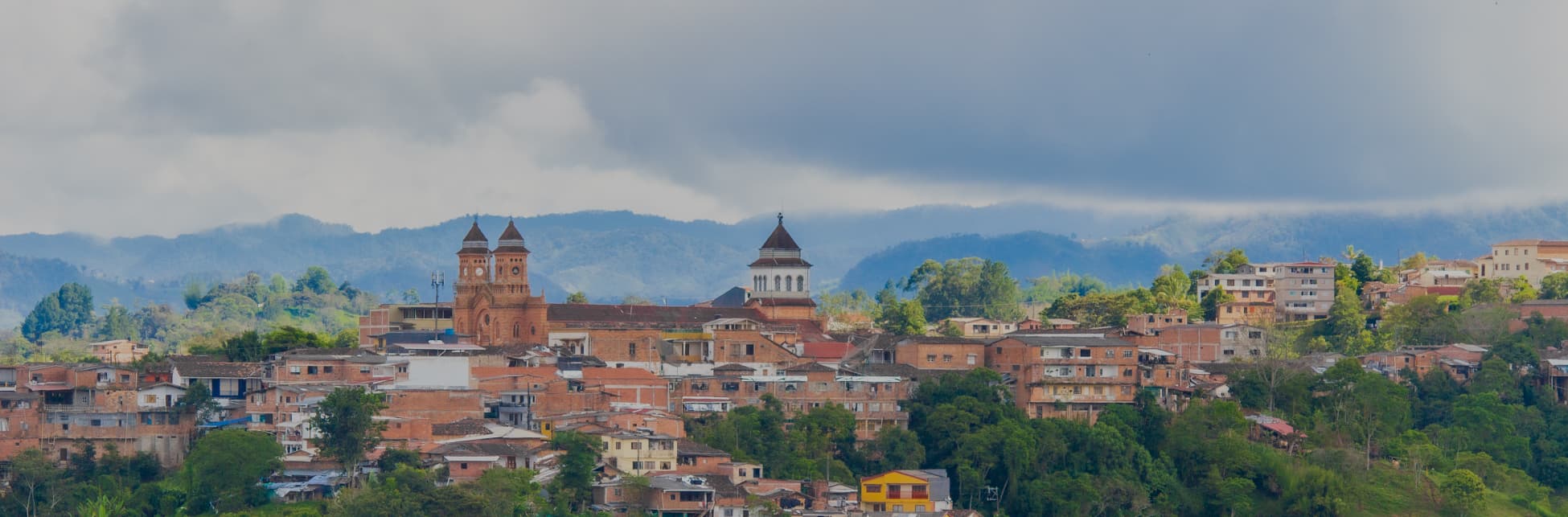Foto. Un grupo de casas u estructuras se erigen alrededor de una iglesia con tes torres. Alrededor hay muchos árboles y al fondo perfiles de montañas. El cielo se encuentra parcialmente nublado.